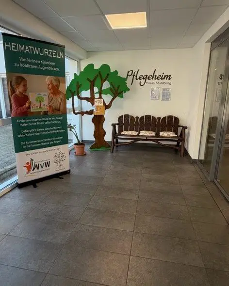 Eingangsbereich eines Pflegeheims mit Sitzbank, bemaltem Baum an der Wand und Roll-up mit Text zu Heimatwurzeln