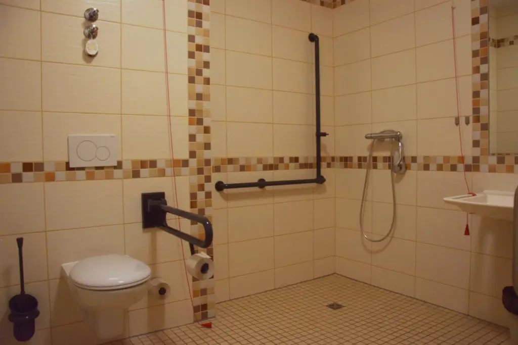 Barrierefreies Badezimmer mit Wandtoilette, Haltegriffen, Duschkopf an der Wand und Waschbecken mit Seifenspender.