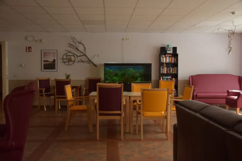 Gemeinschaftsraum mit mehreren Holztischen, Stühlen in Gelb und Braun, Bücherregal, Aquarium und roten Sofas.