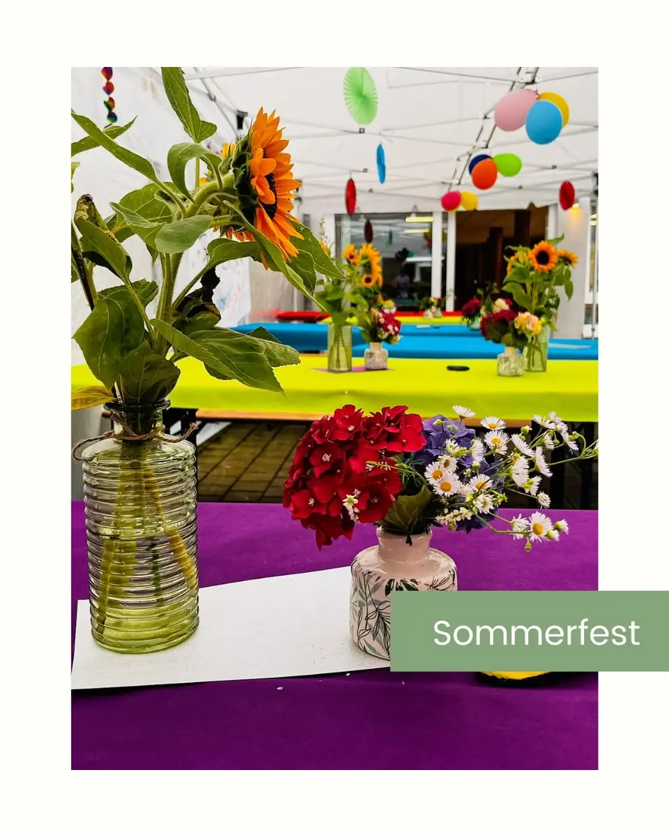 Mehrere bunte Blumensträuße in Glasvasen auf Tischen mit gelben und lila Tischdecken, bunte Papierdekorationen hängen von der Decke.
