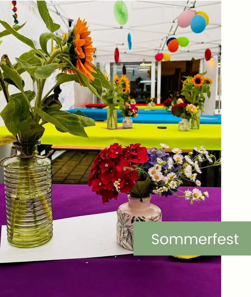 Mehrere bunte Blumensträuße in Glasvasen auf Tischen mit gelben und lila Tischdecken, bunte Papierdekorationen hängen von der Decke.