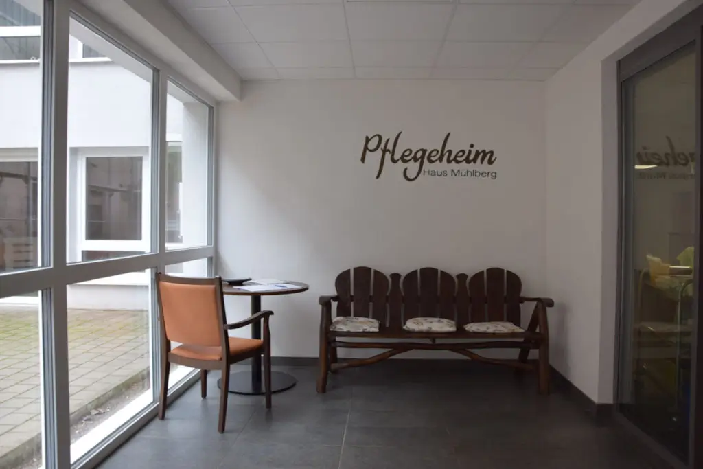 Holzbank mit drei Sitzkissen an weißer Wand mit Schriftzug 'Pflegeheim Haus Mühlberg', daneben runder Tisch mit Stuhl vor großen Fenstern.
