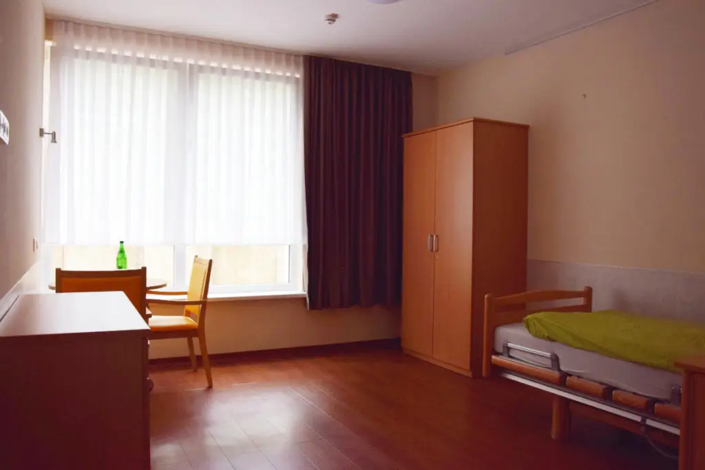 Ein einfach eingerichtetes Zimmer mit Einzelbett, grünem Bettbezug, kleinem Schrank, Schreibtisch und Stuhl vor einem großen Fenster mit halb geschlossenen Vorhängen.