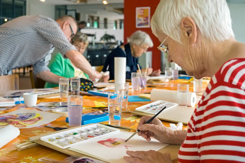 Mehrere ältere Menschen sitzen an einem Tisch und malen mit Aquarellfarben, eine Frau im Vordergrund trägt ein rot-weiß gestreiftes Oberteil