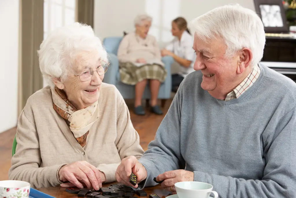 Zwei ältere Menschen sitzen an einem Tisch und spielen Domino, im Hintergrund sitzen zwei weitere Personen auf einem Sessel und einem Stuhl