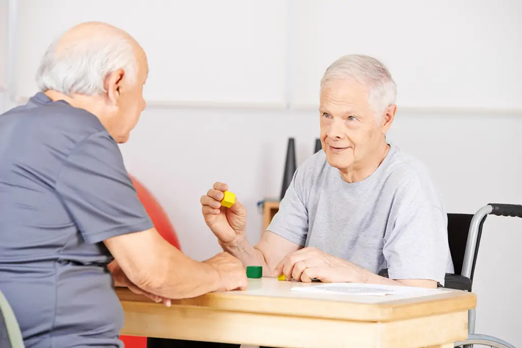 Zwei ältere Männer sitzen an einem Tisch und spielen mit bunten Spielsteinen, einer sitzt im Rollstuhl