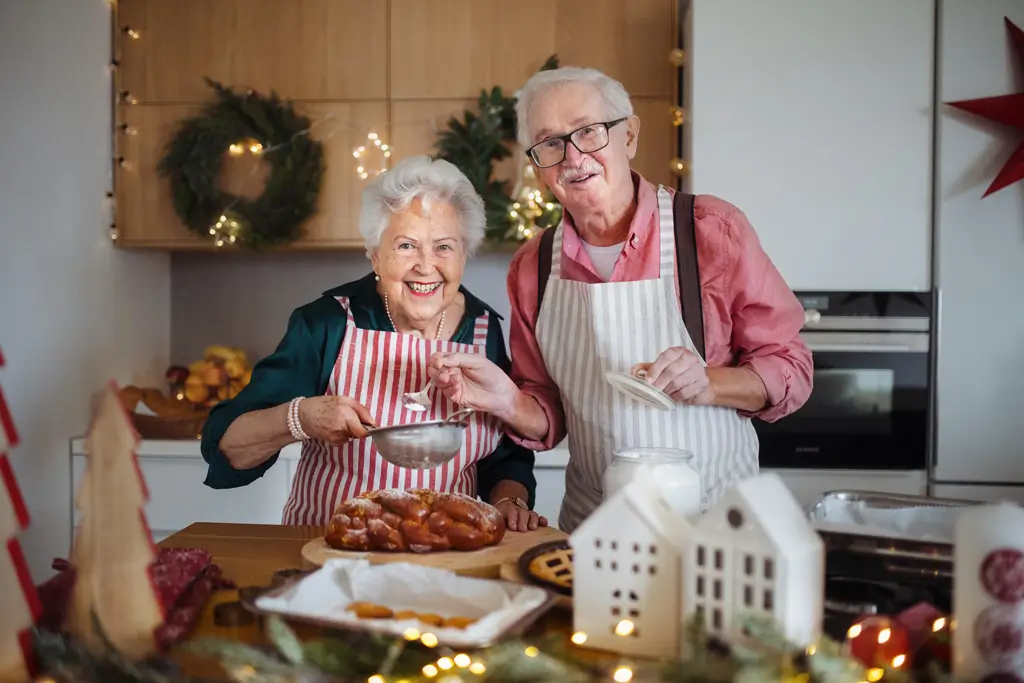 Älteres Paar mit Schürzen in einer weihnachtlich dekorierten Küche beim Backen und Bestäuben von Gebäck